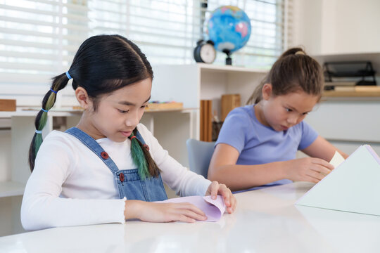 Asian preteen girl folds colored paper carefully while caucasian classmate concentrates nearby classroom desk both enjoying educational origami project improving creativity teamwork and fine motor