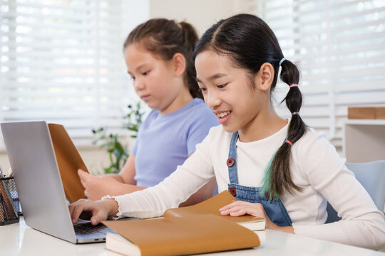 Asian schoolgirl smiling working laptop computer beside caucasian classmate reading notebook desk teamwork classroom project study education learning concept childhood