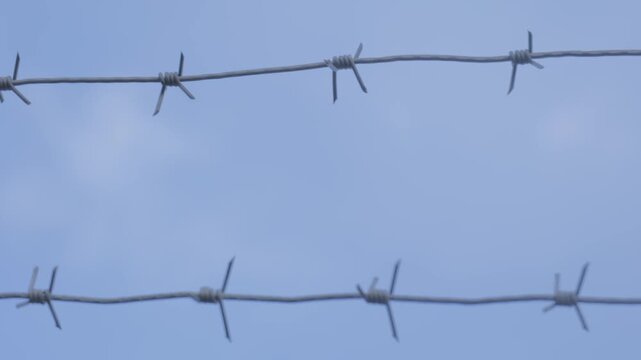 Barbed wire stretches against a blue sky in an open area with some clouds visible on a sunny day