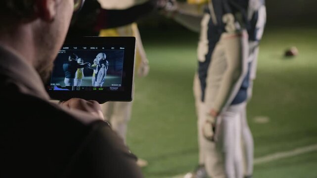 Medium over-shoulder shot of skilled male videographer filming amateur American football team joining hands, cheering for motivation, running off for practice game on pitch, focus on camera viewfinder