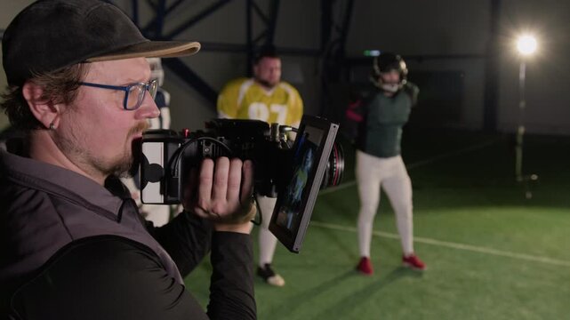 Side medium close-up of Caucasian male cinematographer with camera shooting video portrait of American football team captain, bouncing ball, players in uniform standing nearby on pitch