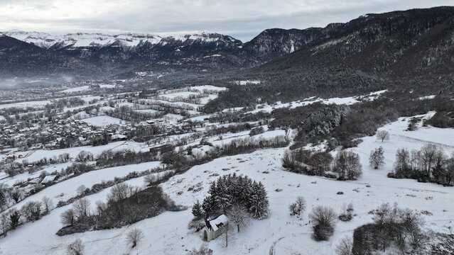 Aerial view of a small chapel nestled among snow-covered trees and fields with the village and Jura mountains in the background Vesancy, Auvergne-Rhone-Alpes, France.