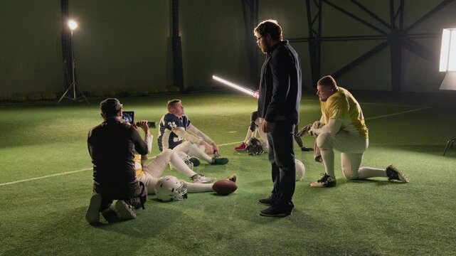 Wide backstage shot of male camera operator filming American football players in gear for commercial, sitting around on green turf at stadium resting during training session, gaffer holding stick lamp