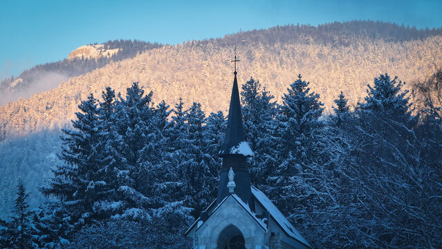 Aerial view of the church of Vesancy spire surrounded by snow-covered pine trees and majestic mountains during winter Vesancy, Auvergne-Rhone-Alpes, France.