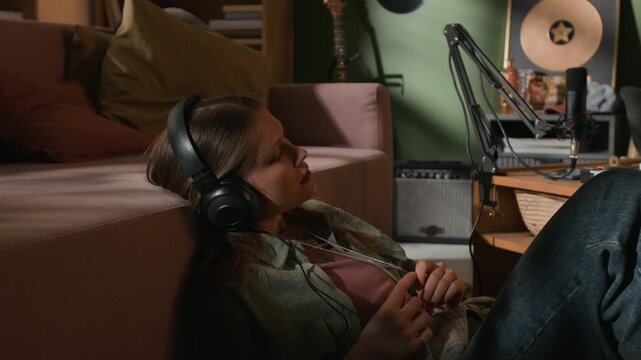 Medium shot of young adult female music producer with headphones on listening to demo track while checking recorded instrumental performance in home studio