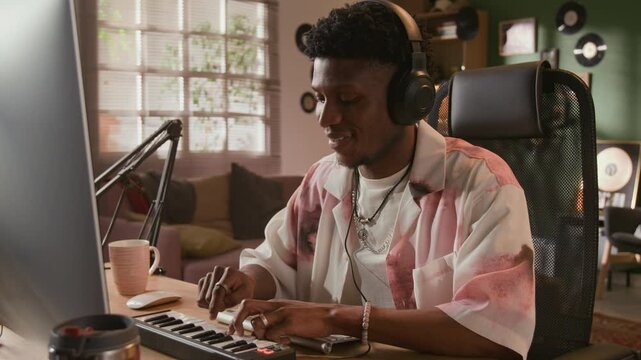 Medium shot of young African American male songwriter with headphones on improvising on MIDI controller keyboard at computer desk while enjoying music production in home recording studio