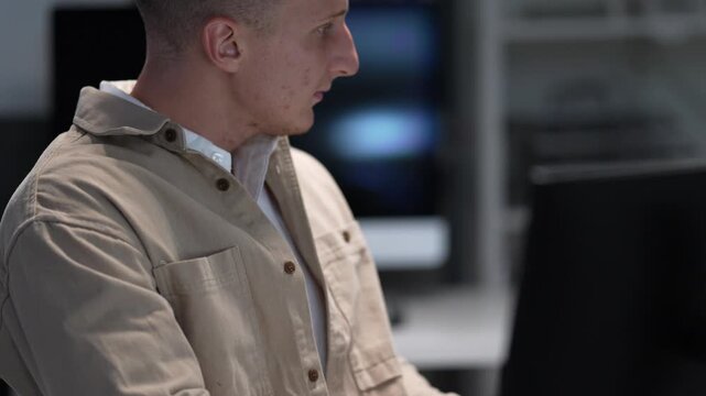 Side profile view of a focused Caucasian tech specialist typing on a computer keyboard while analyzing software interface code at night in a modern office.