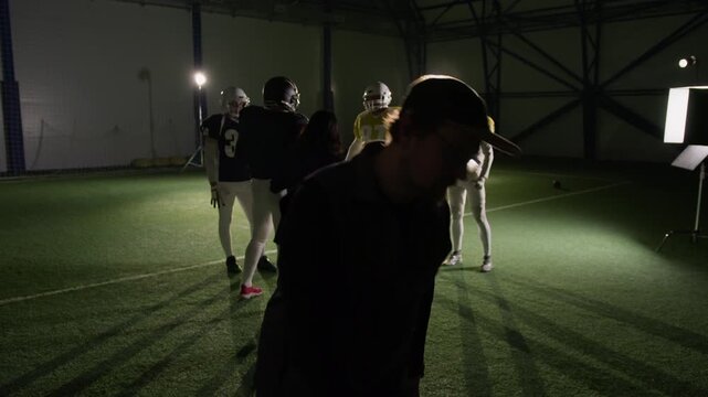 Wide backstage shot of television production crew preparing to film American football team training together at arena, stylist adjusting uniforms while videographer walking to tripod for camera