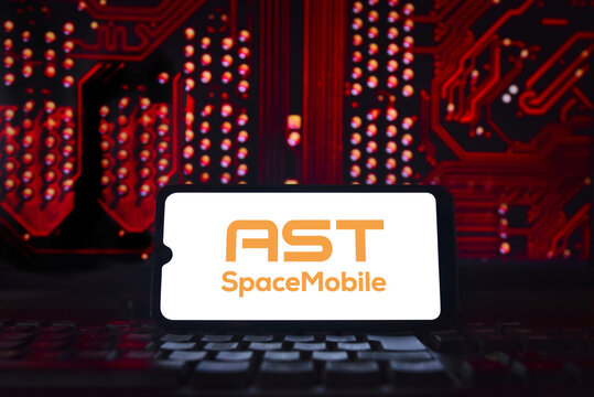 AST SpaceMobile logo displayed on a smartphone screen against a red technological background. The company is developing a cellular broadband network in space to connect smartphones directly to its sat