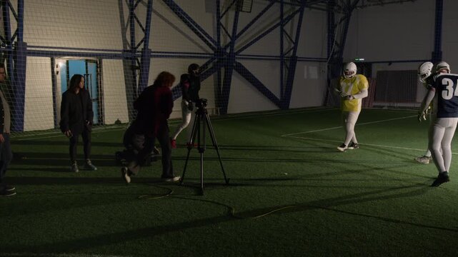 Wide shot of gridiron football players practicing blocking and tackling opponents running with ball during training at sports arena, recorded by professional production crew for television program
