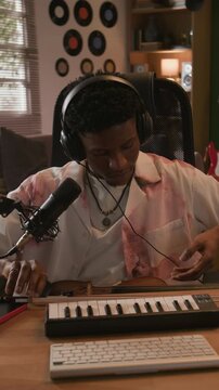 Vertical medium shot of young adult Black male musician putting on headphones before recording violin performance at desk with MIDI controller in home studio