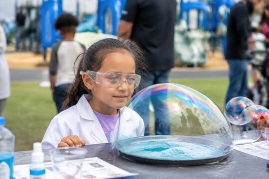 A young girl in a lab coat and safety goggles observes a large iridescent bubble during an outdoor science event. Captures the joy of learning and curiosity for educational use.