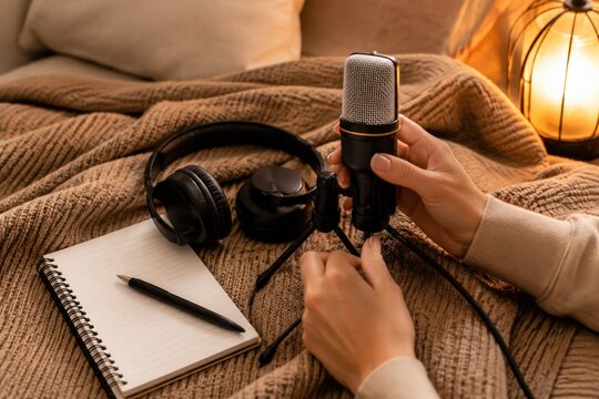 Close up of hands adjusting a professional microphone on a cozy bed with headphones and notebook. Warm atmosphere for content creation. Ideal for podcasting and media production ads.