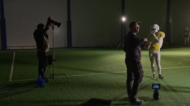 Wide panning shot of Caucasian male videographer discussing scene for shooting with gridiron football team for TV sports reality show, and instructing gaffer to reposition lights at arena