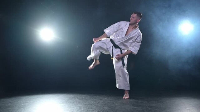 Strong man fighter in white kimono for karate martial arts with black belt preparing for fight training in black background. Man training moves punches kicks practicing shadow fight ready for sparring