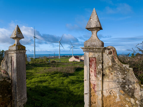 Aerial view of historic stone gate pillars overlooking a green field with wind turbines and the ocean in the distance in Ribamar, Portugal.