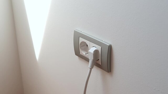 Closeup of a hand plugging a cord into a wall outlet, showing a clear electrical connection in a plain indoor setting.