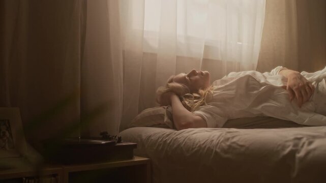 Handheld shot of blonde Caucasian young woman relaxing in bed and enjoying music on vinyl record player in cozy home morning