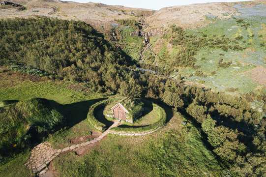 Aerial view of The Commonwealth Farm turf house with its circular stone wall surrounded by lush green hills and a distant waterfall in Selfoss, Skeida- og Gnupverjah, Iceland.