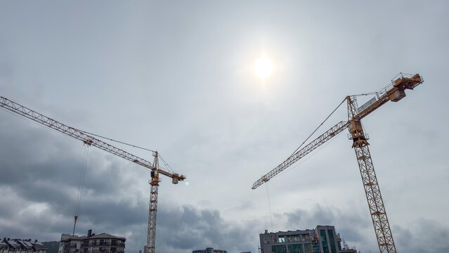 Construction cranes against a cloudy sky with sun peeking through clouds