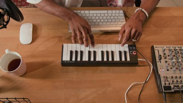 Top view close up of unrecognizable Black male music producer playing MIDI controller keyboard at production setup equipped with mixing console while recording electronic track in home studio