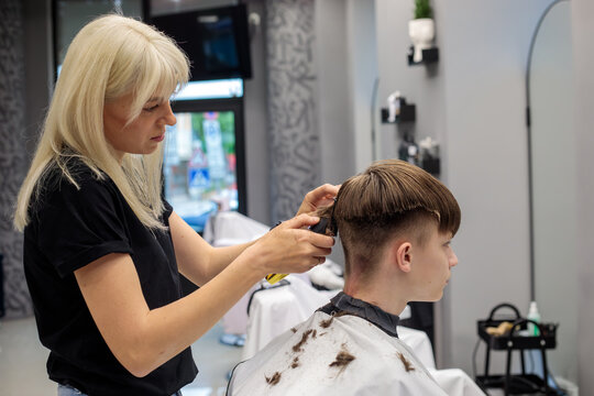 wide shot of blonde female barber using clipper to cut teenage boy hair in modern barbershop with mirror grey wall and styling tools visible
