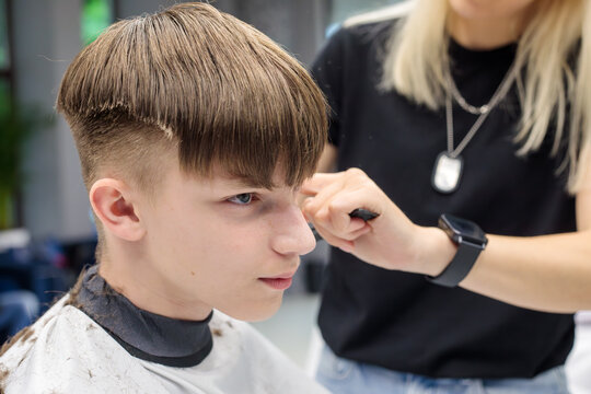 portrait of teenage boy with completed bowl cut low fade while blonde barber holds trimmer near his temple in modern barbershop interior