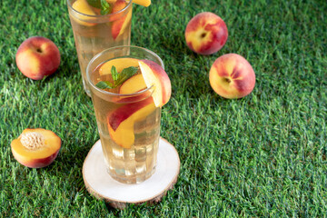 Refreshing glasses of homemade peach iced tea with mint leaves and fresh fruits scattered on a vibrant green lawn © Magdalena