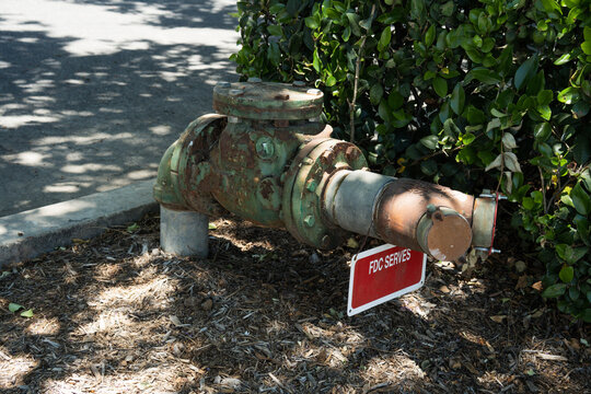 Old rusty fire department connection valve with red sign in front of green bushes