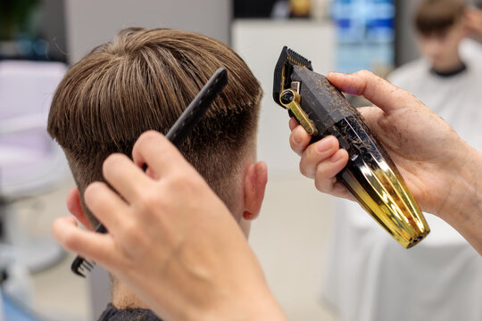 back view of teenage boy head with fade haircut while barber holds gold trimmer in one hand and black comb in other at barbershop