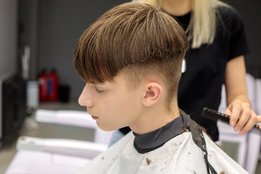  side profile of teenage boy with bowl cut fade haircut looking down while blonde female barber holds comb near neck in grey barbershop