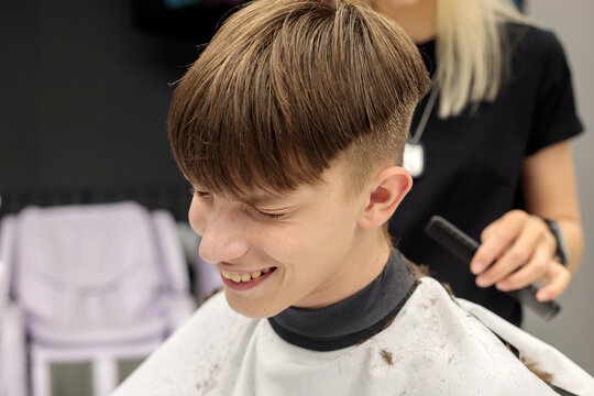 happy smiling teenage boy with fresh bowl cut fade haircut looking down while blonde female barber combs hair from behind in barbershop