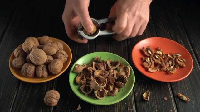 Hands cracking open walnuts using a nutcracker, with shells and nuts arranged on colorful plates against a dark wooden background