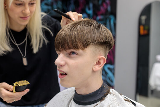 blonde female barber using comb and gold trimmer to style teenage boy bowl cut while boy talks with mouth open in modern barbershop
