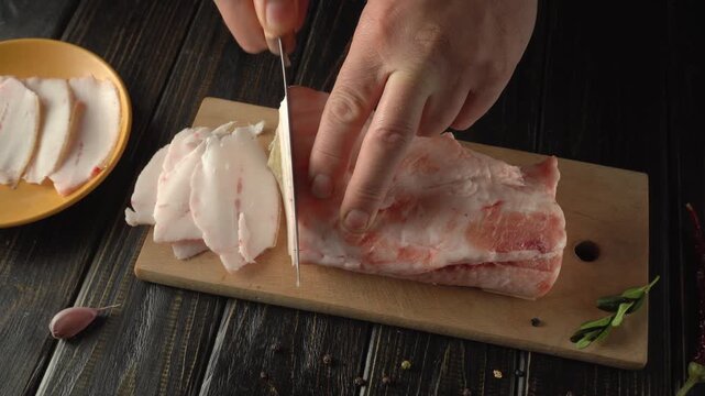 Hands of a Caucasian male chef slicing pork fat on a wooden cutting board with a yellow plate and garlic visible in the background
