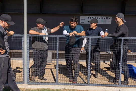 Five men in athletic gear leaning on chainlink fence at dugout scoreboard holding water bottles