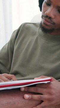 Vertical video: Glancing left, man flipping red notebook and writing notes with pen at home