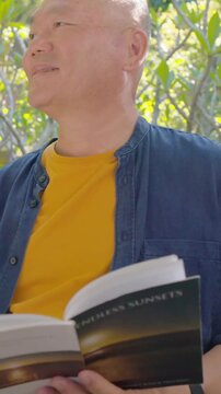 Vertical video: Hearing birds, senior Asian man turning book pages on patio bench to relax, smiling