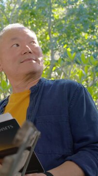 Vertical video: Holding book, Asian man in yellow tee checking offscreen, resuming reading in park