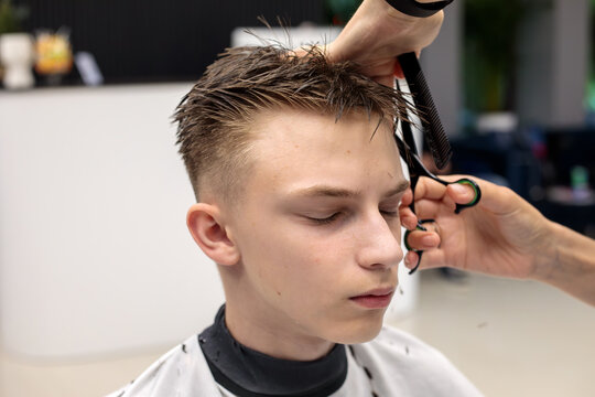 barber using scissors and comb to cut wet top hair of teenage boy with high fade haircut in modern bright barbershop setting