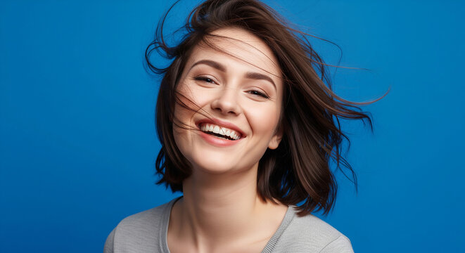 Joyful woman laughing with her hair blowing in the wind on a blue background, perfect for carefree and happy beauty concepts