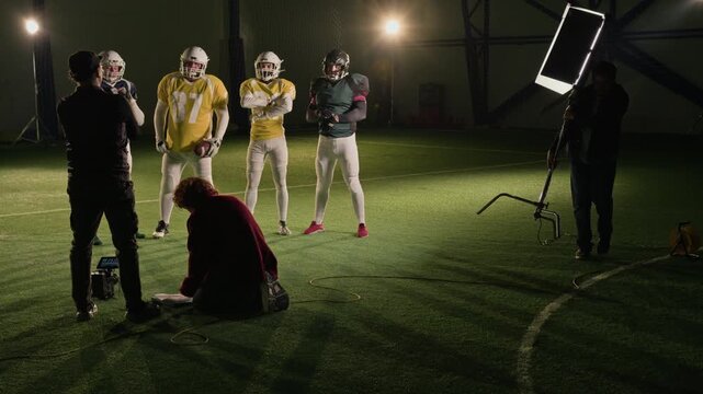 Wide behind scenes shot of TV production crew preparing to film group portrait of gridiron football team in match gear, standing on pitch, camera operator directing position while gaffer moving lights