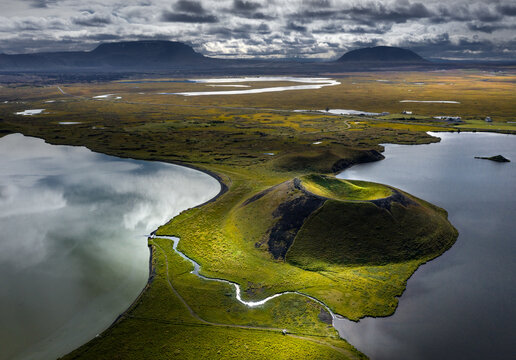 Aerial view of the Skutustadagigar pseudocraters at Lake Myvatn surrounded by green mossy landscape and calm water under a dramatic sky in Iceland.