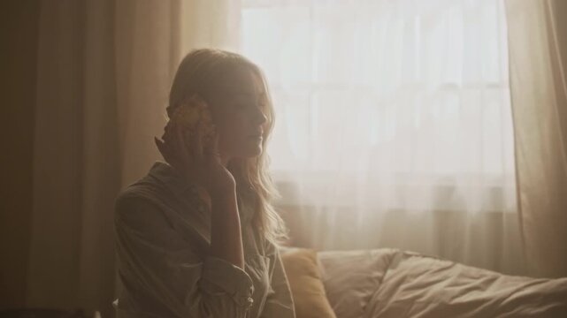 Zoom in shot of delicate young woman listening to sound from seashell and reminiscing about ocean travel while resting on bed under morning light