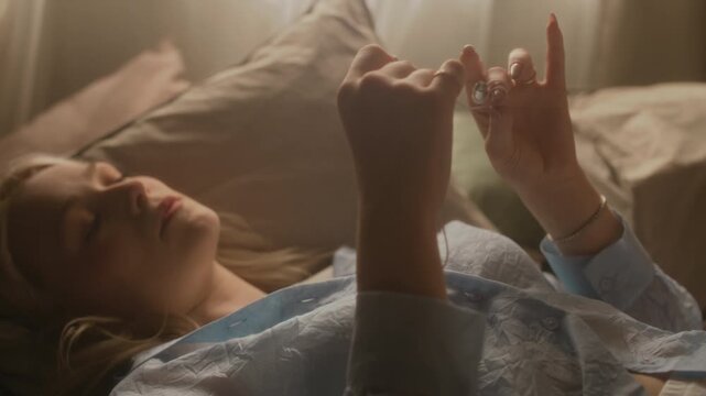 Handheld slow motion of dreamy gen Z girl lying in bed under morning sunlight, twirling wired earphones around fingers, lost in thought