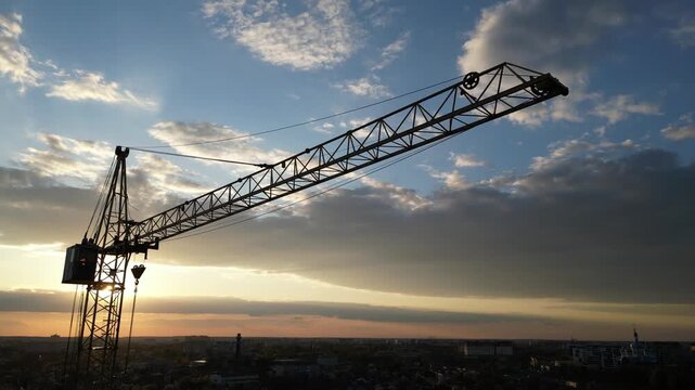 Majestic aerial view showing a large construction tower crane descending against a beautiful sunset sky, with the sun's rays creating a stunning lens flare over a darkening cityscape