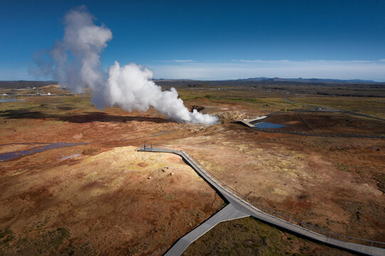 Aerial view of a geothermal area with a massive steam plume rising from a vent, wooden walkways, and people on a viewing platform, Iceland.