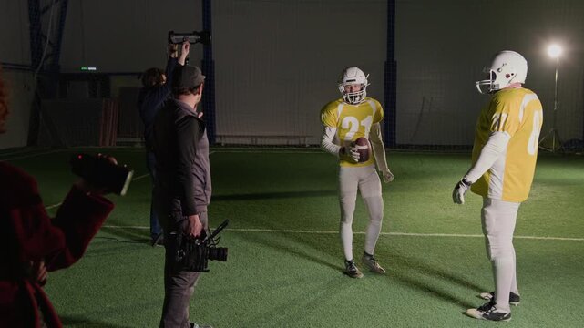 Full shot of television production crew setting up scene for shooting American football TV program, with gaffer positioning light, camera operator talking to sportsmen and producer observing action