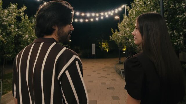Young couple walks hand in hand through park with blooming trees before sharing kiss. Romantic night stroll creates warm intimate atmosphere.