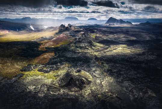 Aerial view of the rugged volcanic landscape featuring dark lava fields, mossy craters, and geothermal steam under a dramatic sky with light rays Iceland.